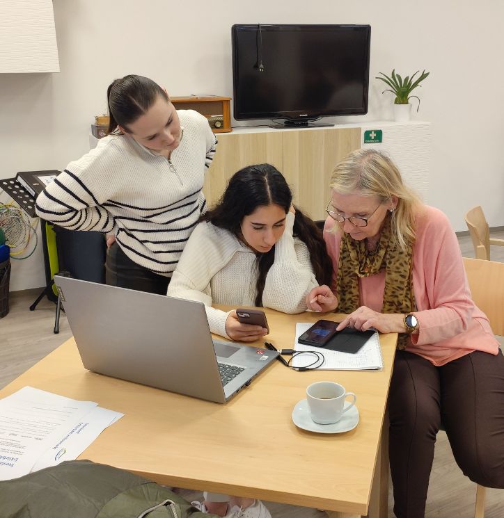 Eine junge Frau erklären einer älteren Dame etwas auf dem Smartphone. Beide sitzen an einem Tisch, vor ihnen ein Laptop. Eine weitere junge Frau steht hinter ihnen und seht ihnen über die Schulter. © Caritas Salzburg
