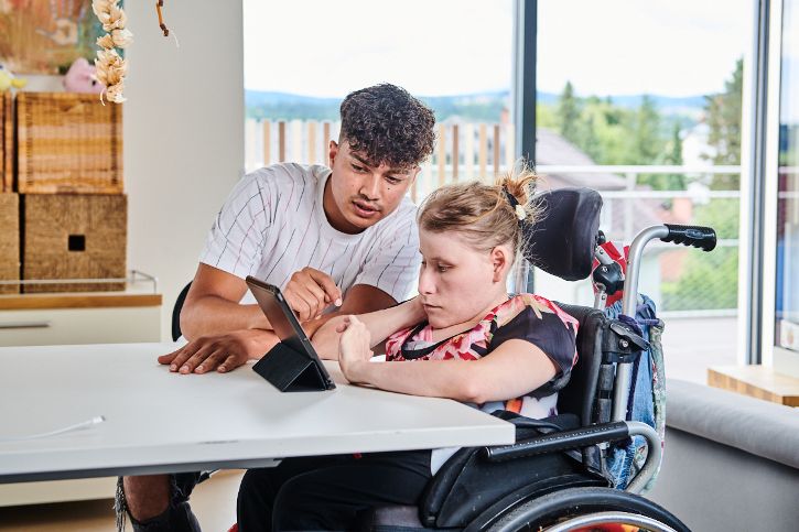 Ein junger Mann erklärt einer Frau im Rollstuhl etwas auf einem Tablet. © Christopher Mavric - LebensGroß