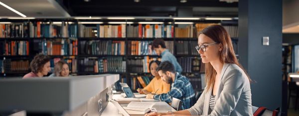 Universitätsbibliothek: Talentiertes kaukasisches Mädchen sitzt am Schreibtisch, benutzt Laptop, schreibt Notizen für die Hausarbeit, Aufsätze, lernt für die Klassenaufgabe. Vielfältige Gruppe von Studenten lernt, lernt für Prüfungen.