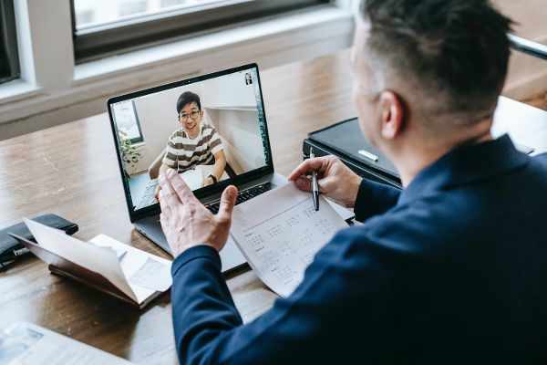 Ein Mann mit dem Rücken zur Kamera an einem Tisch sitzend. Vor ihm ein Laptop mit einer aktiven Videokonferenz.