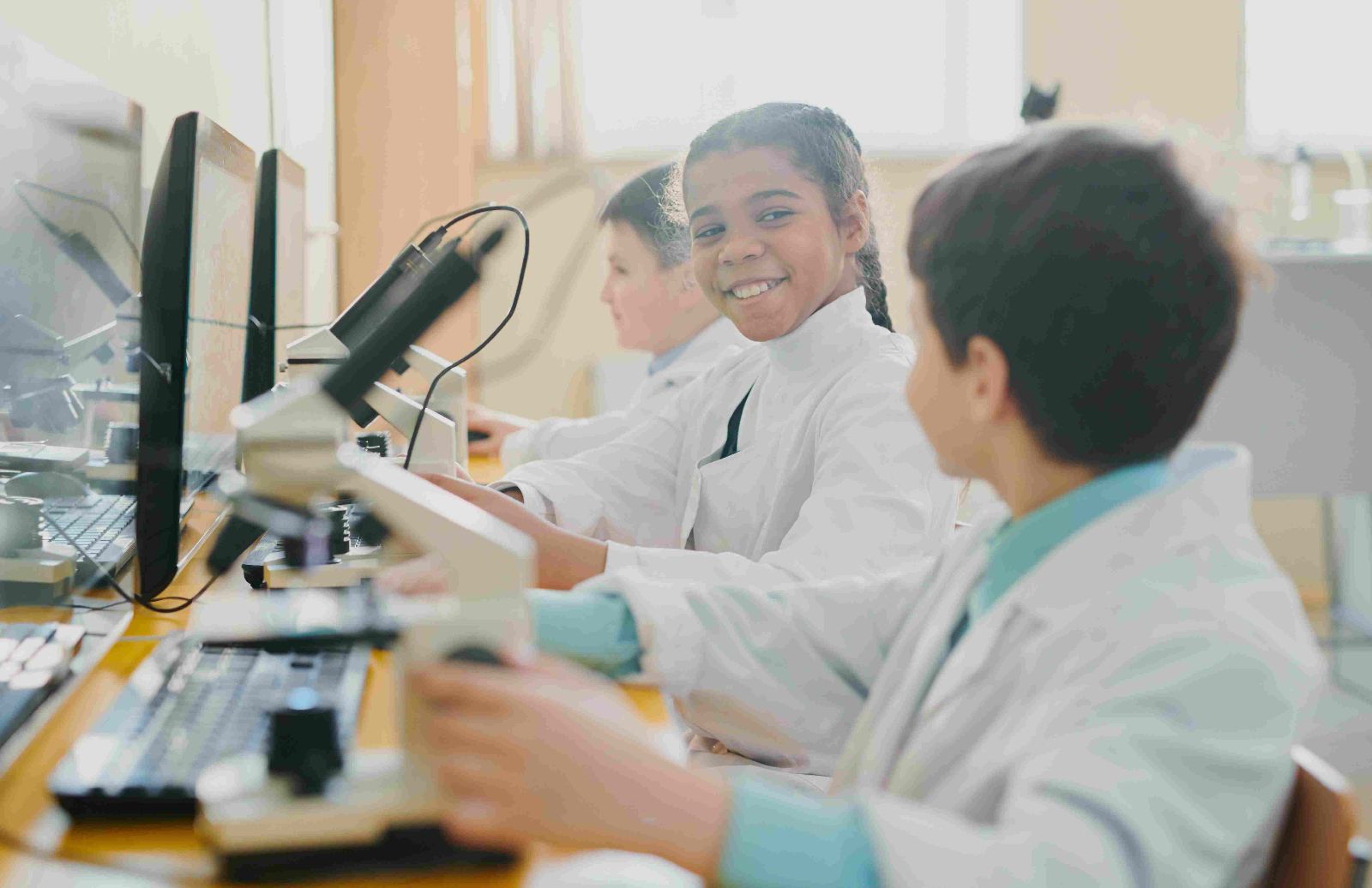 Drei Kinder, die in Laboruniform vor einem Computer und Mikroskop sitzen © Unsplash+