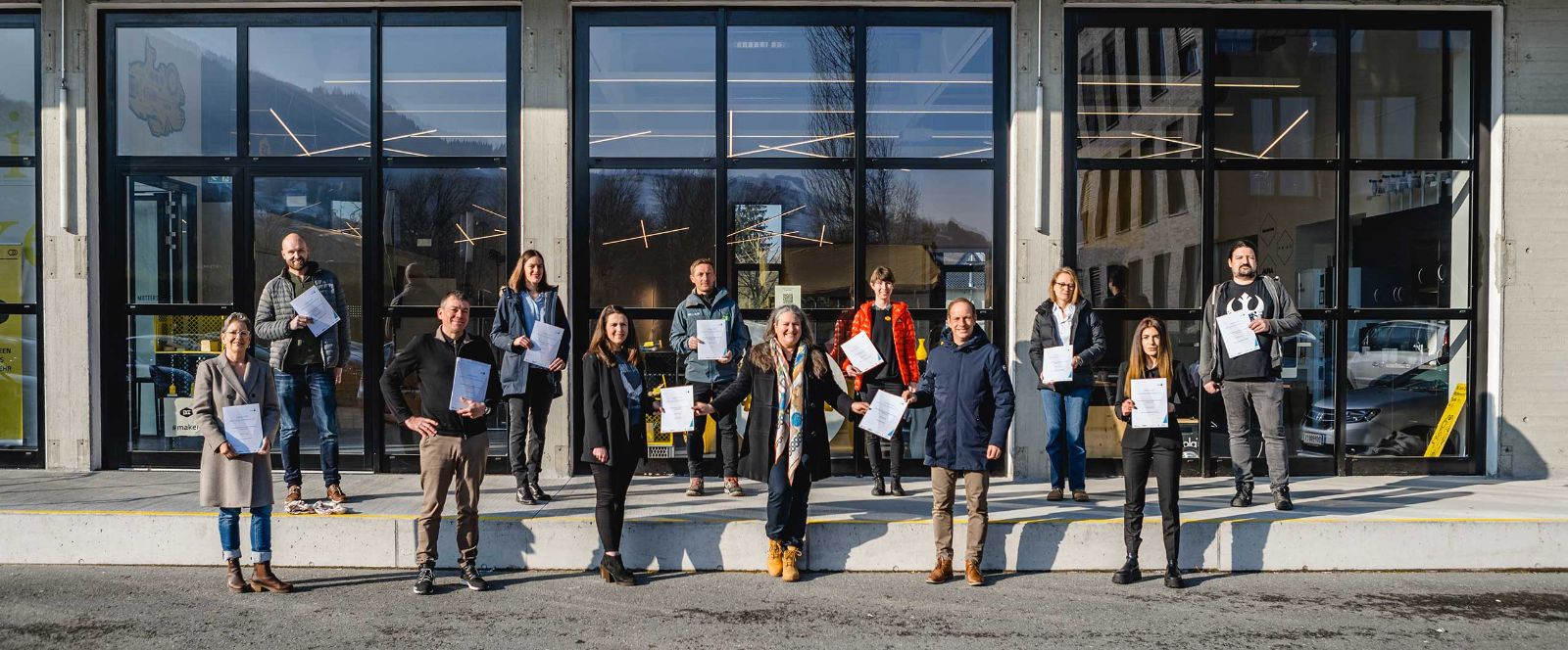 Eine Gruppe Menschen steht mit jeweils einem Ordner in der Hand vor einem großen, gläsernen Gebäude. © Mint4all Vorarlberg - Mint4all