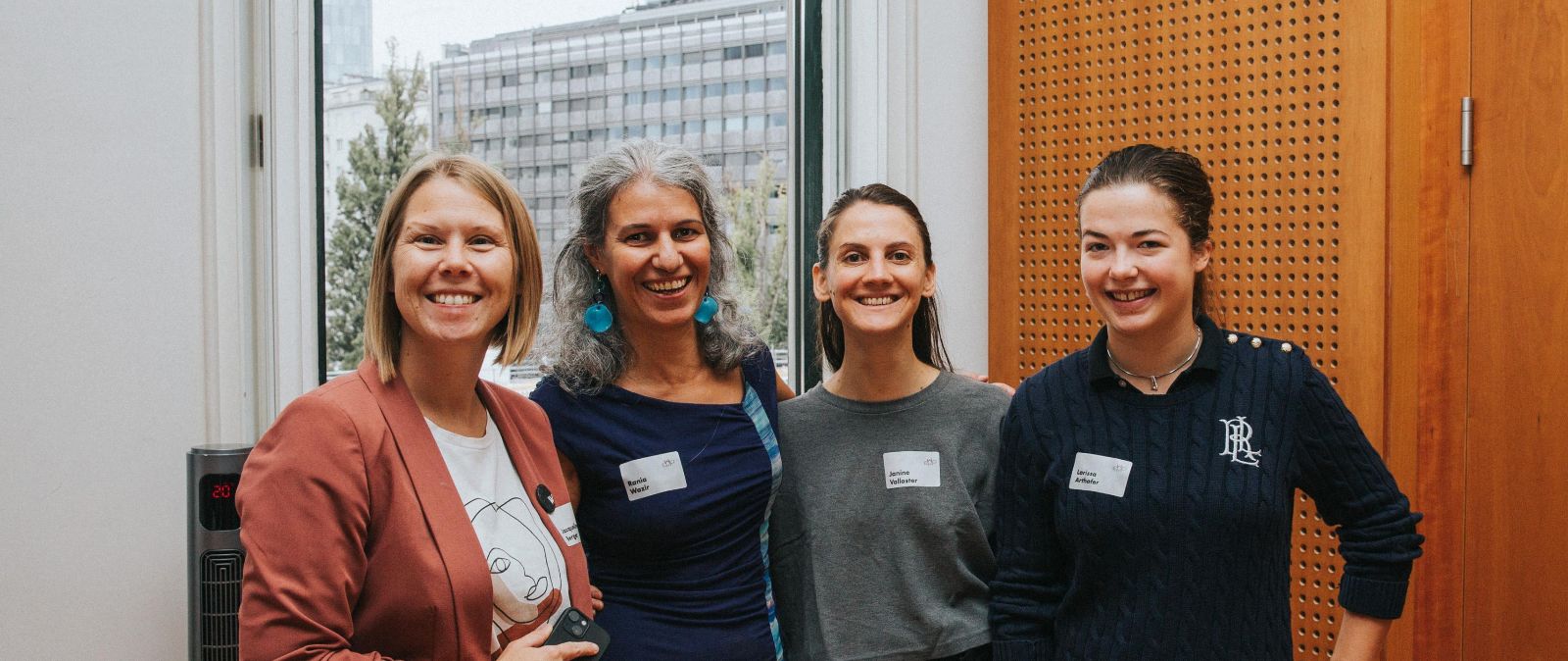 Zu sehen sind vier Frauen, die gemeinsam in die Kamera lächeln. Im Hintergrund ist ein großes Fenster zu sehen und auf der rechten Seite große Holz-Paneele. © Kathrin Hanga - Women in AI Austria
