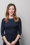 Zu sehen ist eine junge Frau mit langen, braunen Haaren. Sie lächelt in die Kamera und trägt ein dunkelblaues Kleid.