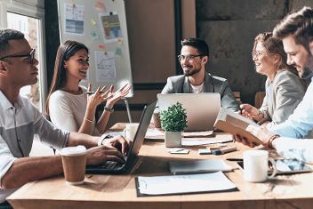 Gruppe junger Menschen sitzen sich an einem Tisch mit geschäftlichen Unterlagen und aufgeklappten Laptops gegenüber. &copy;  Adobe Stock 