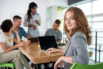An einem Schreibtisch sitzende Frau, mit Laptop vor sich, schaut direkt in die Kamera. Ebenfalls beim Tisch sitzen vier weitere, in ein Gespräch vertiefte Personen. &copy;  Adobe Stock 