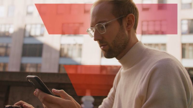 Mann mit Brille sitzt vor einem Gebäude und hält ein Smartphone in der Hand. © Monte Nero Productions  - Monte Nero Productions 