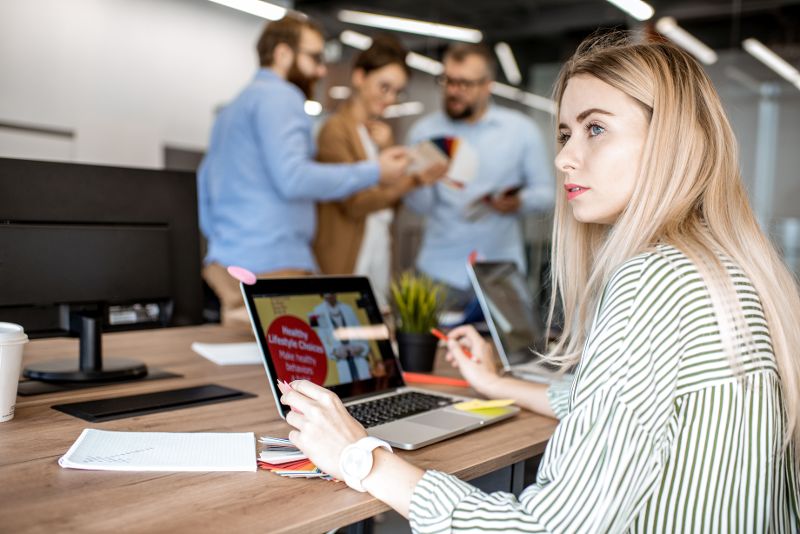 Eine Frau sitzt vor einem Laptop und blickt in die Ferne. Im Hintergrund stehen drei Personen beisammen.