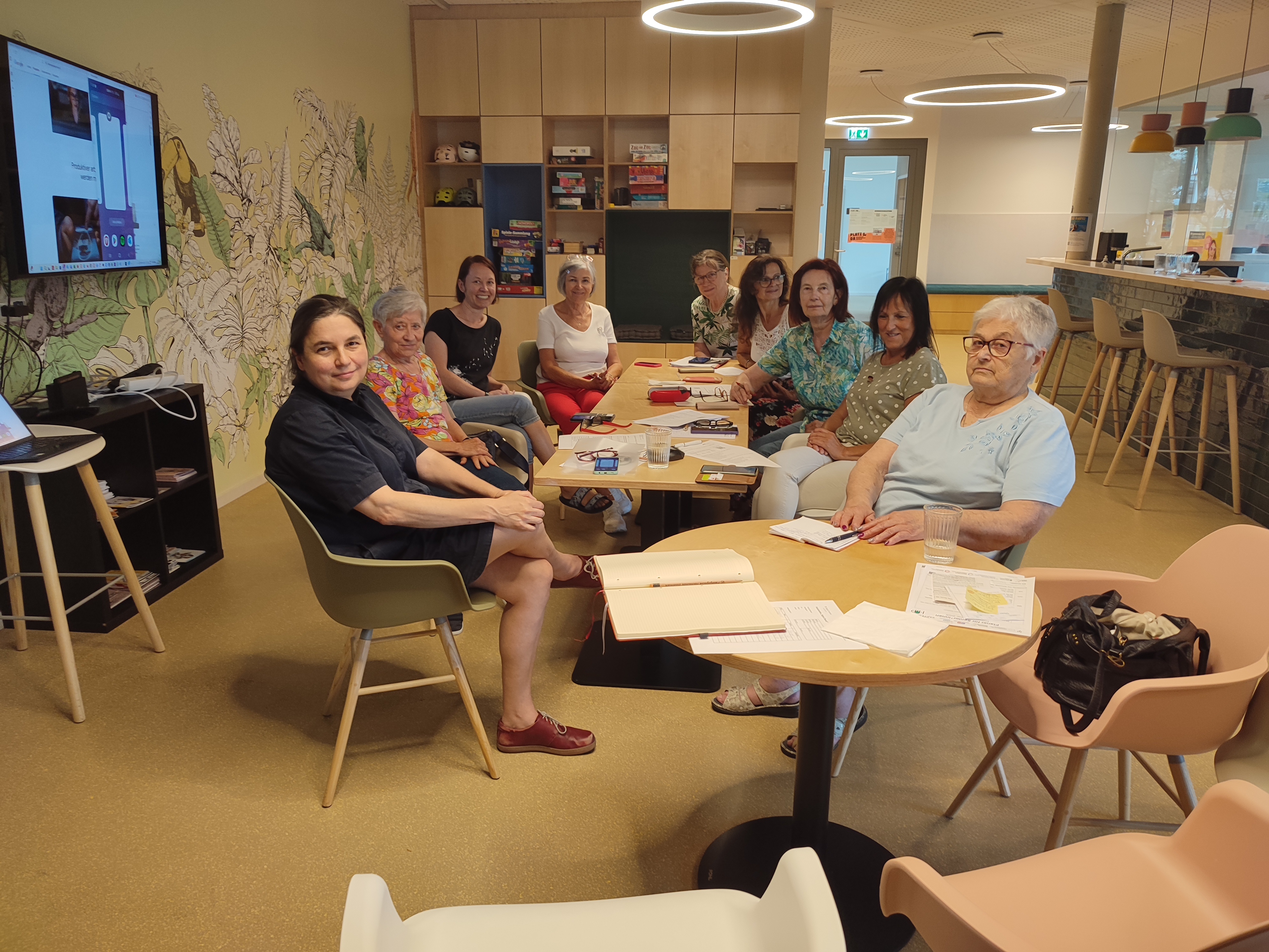 Foto von einem Digital Überall Workshop auf dem Seniorinnen rund um einen Tisch sitzen und in die Kamera schauen.